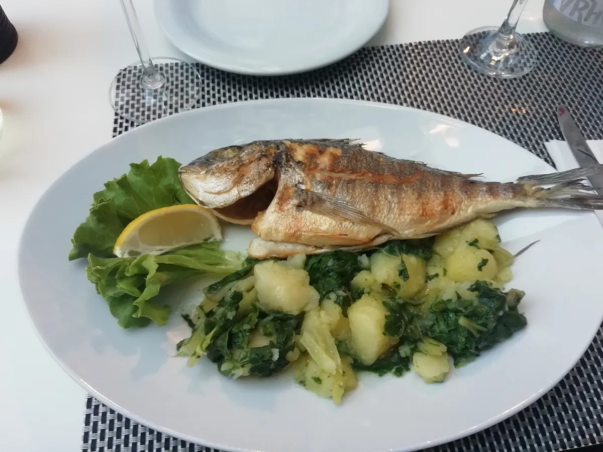 Whole grilled fish with a lemon wedge, lettuce and potatoes with greens on a white plate