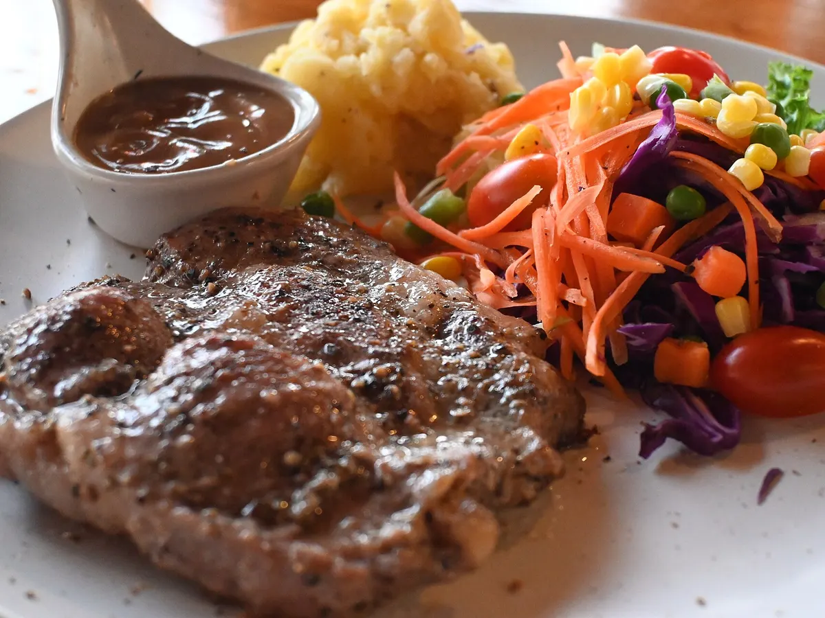Grilled beef steak on a white plate with a jug of pepper gravy, mashed potatoes and a colourful vegetable salad